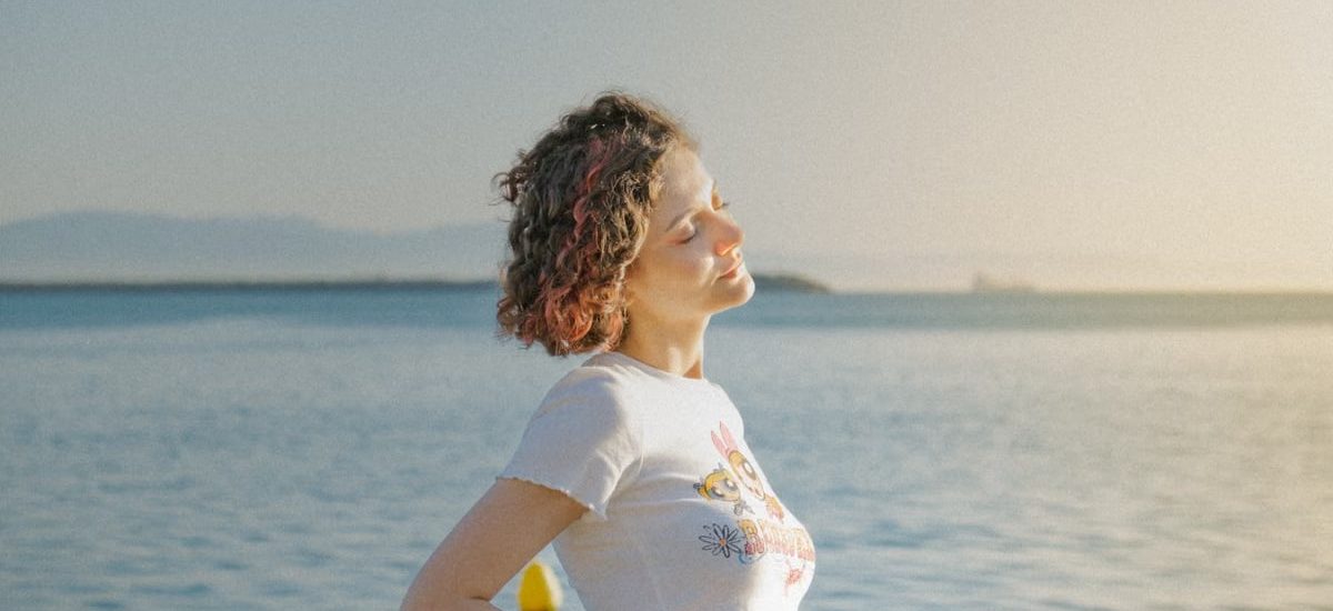 A woman with eyes closed stands by the ocean, embracing the sunlight and tranquility.