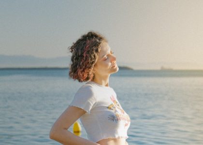 A woman with eyes closed stands by the ocean, embracing the sunlight and tranquility.